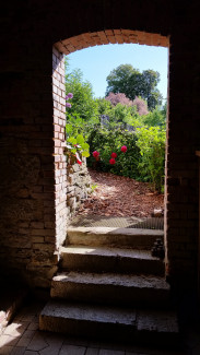 Dunkle Stufen führen durch eine Tür in einer dunklen Mauer. Dahinter ist ein blühender Garten zu sehen.